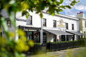 White two-story building housing 'The Beehive' pub, with gold lettering, black awnings, and a 'Montpelier Villas' sign, framed by leafy foreground.