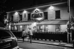 Nighttime street scene of The Beehive pub with illuminated sign and awning; blurred pedestrians in foreground.