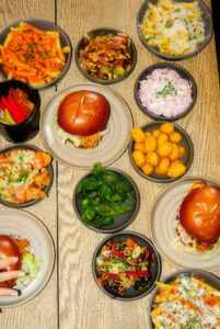 Assorted bowls of food on a wooden table, including burgers, vegetables, and grains, ready for sharing