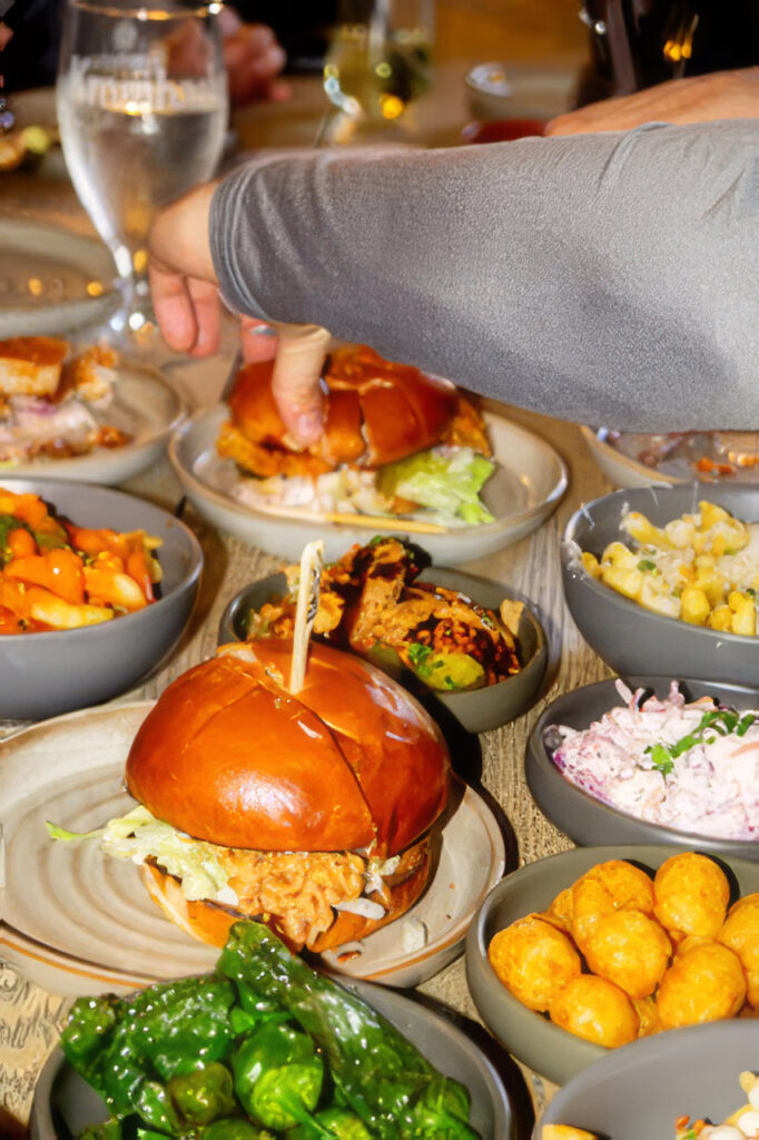 Arm reaching for a glossy hamburger at a table filled with bowls of assorted side dishes.