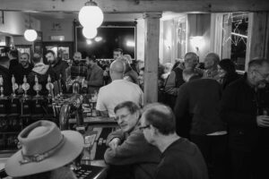 Crowded bar with patrons chatting and drinking; beer taps run along the counter at the foreground.