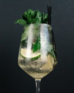 Glassy cocktail with lime wedges, mint leaves, ice, and a black straw on a dark background (sparkling drink).