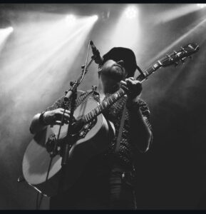 Male guitarist wearing a hat performs on stage, singing into a microphone while playing an acoustic guitar under bright stage lights (black-and-white photo).
