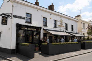 White storefronts of two-story pubs with 'The Beehive' signage and outdoor seating, people chatting along the sidewalk under awnings, blue sky above.