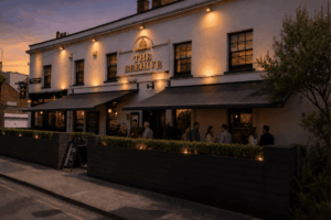 Exterior of The Beehive pub at dusk with warm outdoor lights and groups of people standing and chatting outside.