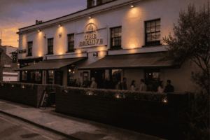 Exterior of a two-story pub named 'The Beehive' with warm wall lights and outdoor seating at dusk, people socializing outside