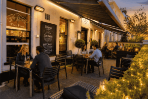 Outdoor restaurant patio at dusk with groups of people dining under warm string lights and lanterns.